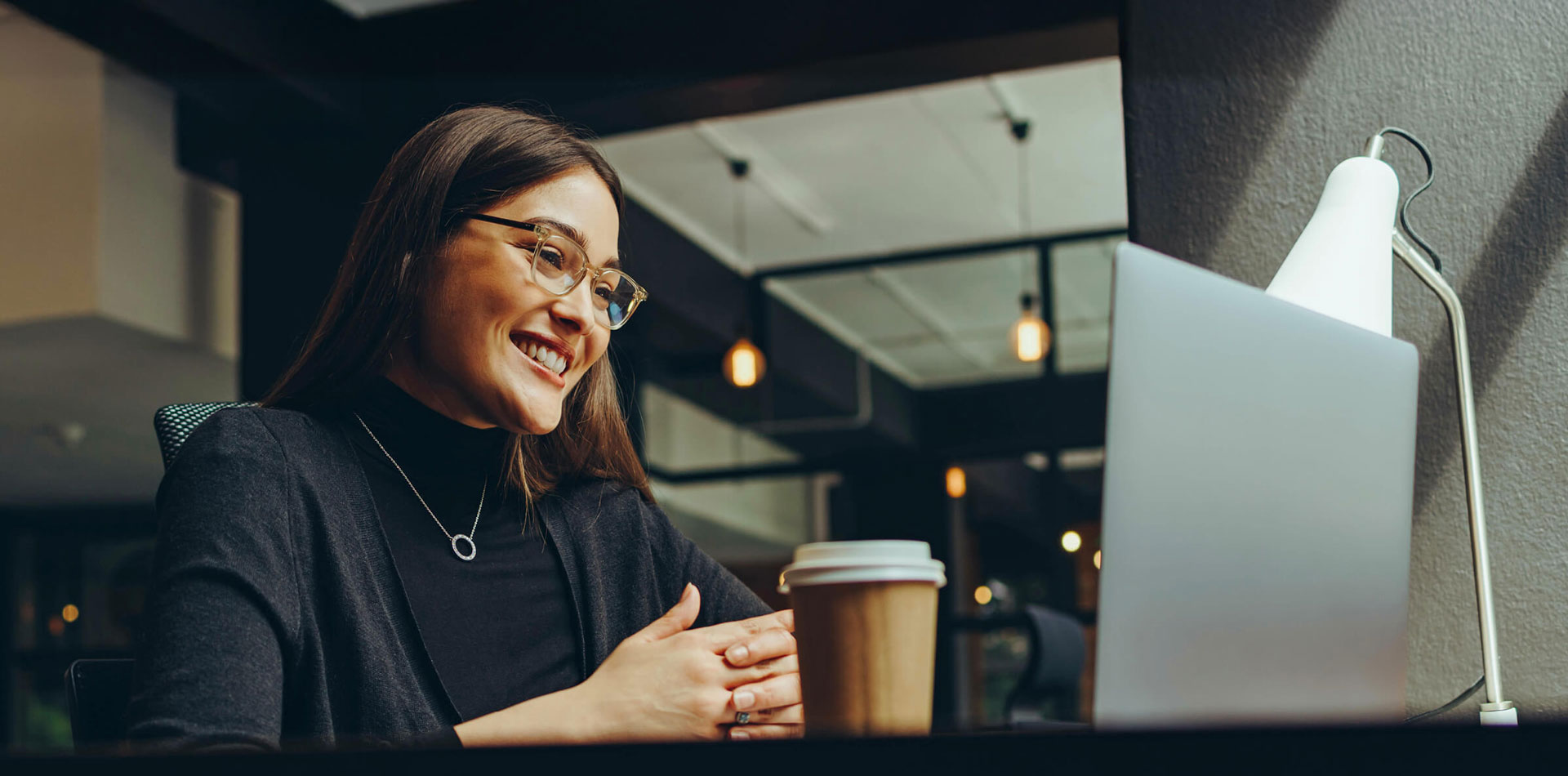 A smiling woman in glasses and dark clothing is seated at a table, looking at a laptop with a coffee cup and lamp beside it.