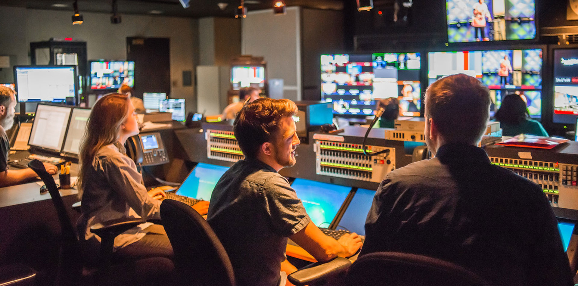 A group of people working in a dimly lit TV control room with multiple screens and equipment.