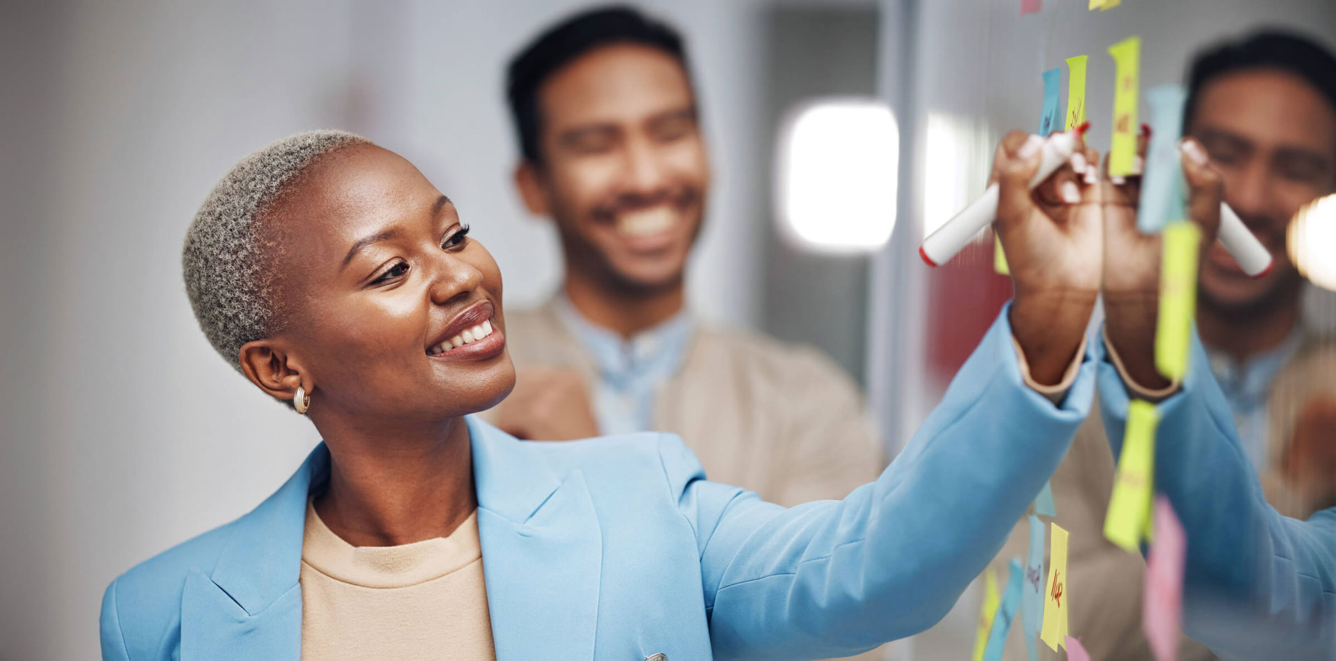 A smiling woman writes on a glass wall covered with sticky notes, with a colleague nearby.